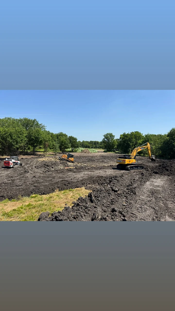 Excavator and skid steer loader grading dark soil on cleared land with trees in background