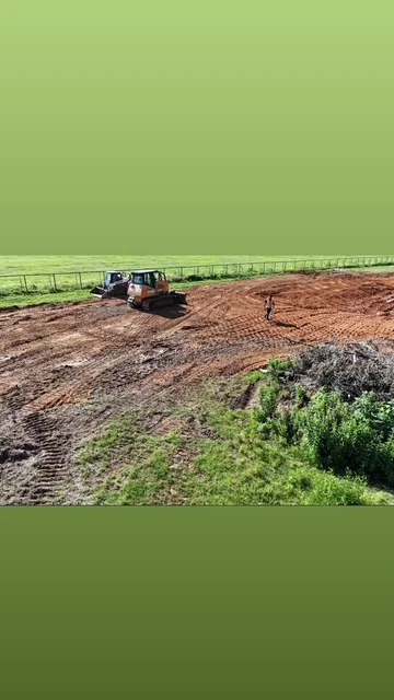 Tracked excavator grading and leveling brown soil on large property with green pasture beyond