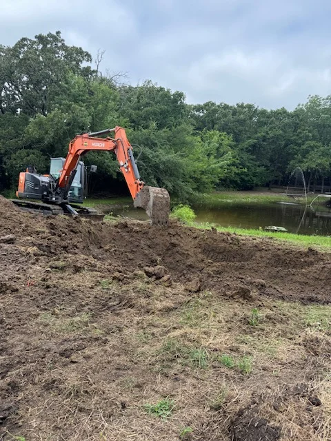 Orange tracked excavator moving soil on waterfront property with trees and water visible
