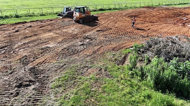 Tracked excavator grading brown soil on cleared land with green vegetation borders