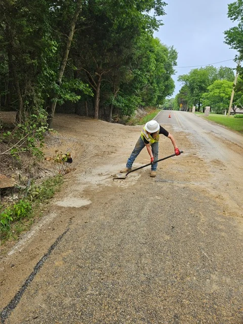 Worker grading and compacting driveway base material with hand tools on rural road