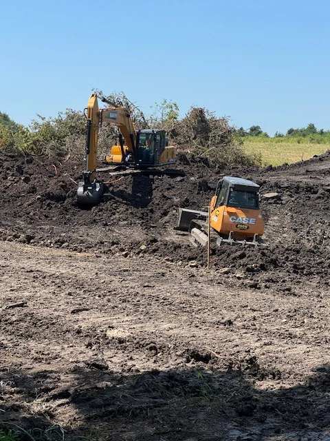 Excavator clearing brush and vegetation with support truck on prepared dirt site