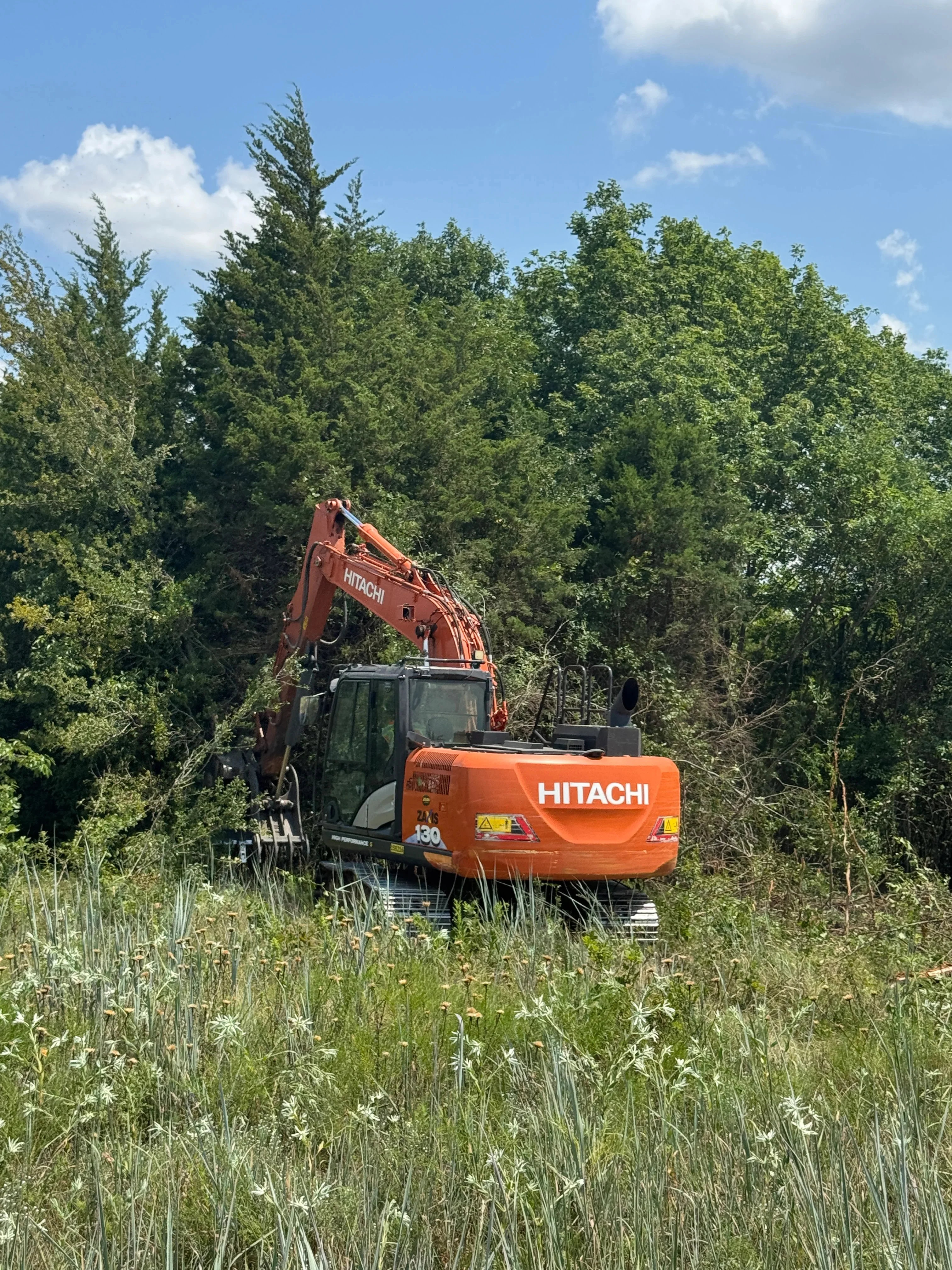 A Dallas property lot after professional brush removal, showing cleared ground with cedar and mesquite stumps cut to grade and open land ready for use. A Patriot Equipment and Landworks crew member operates a forestry mulcher along a fence line with native oak trees preserved in the background.