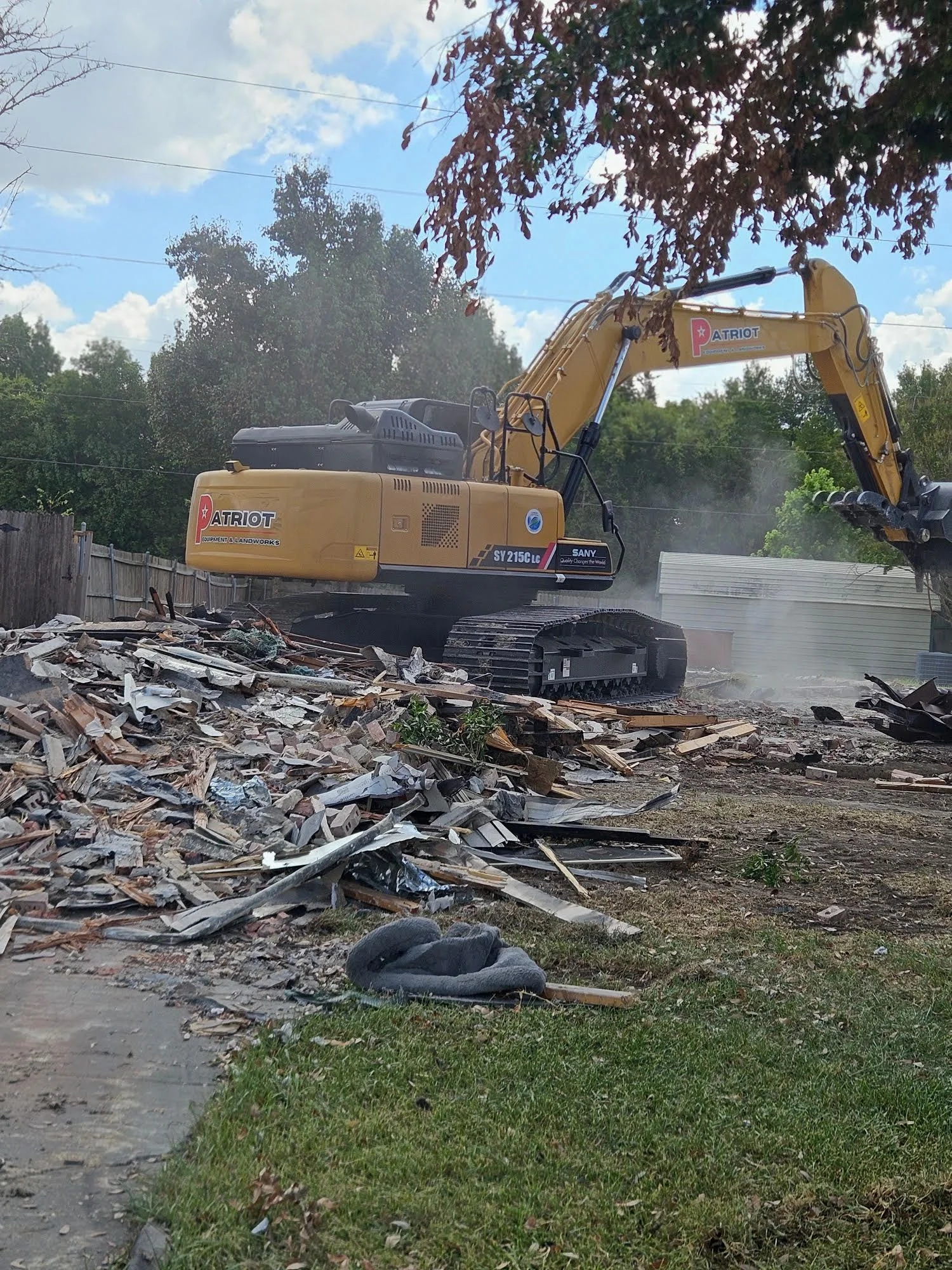 A tracked excavator breaking down a single-story residential structure on a cleared Dallas lot, with debris staged for removal and surrounding properties undisturbed. The site shows a full building demolition in progress under a clear Texas sky.