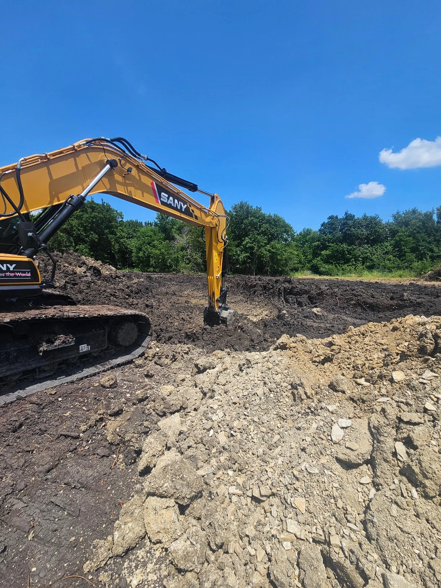 A tracked excavator operated by Patriot Equipment and Landworks digging a foundation cut on a cleared residential lot in Dallas, Texas. Exposed clay soil and a graded building pad are visible in the foreground.