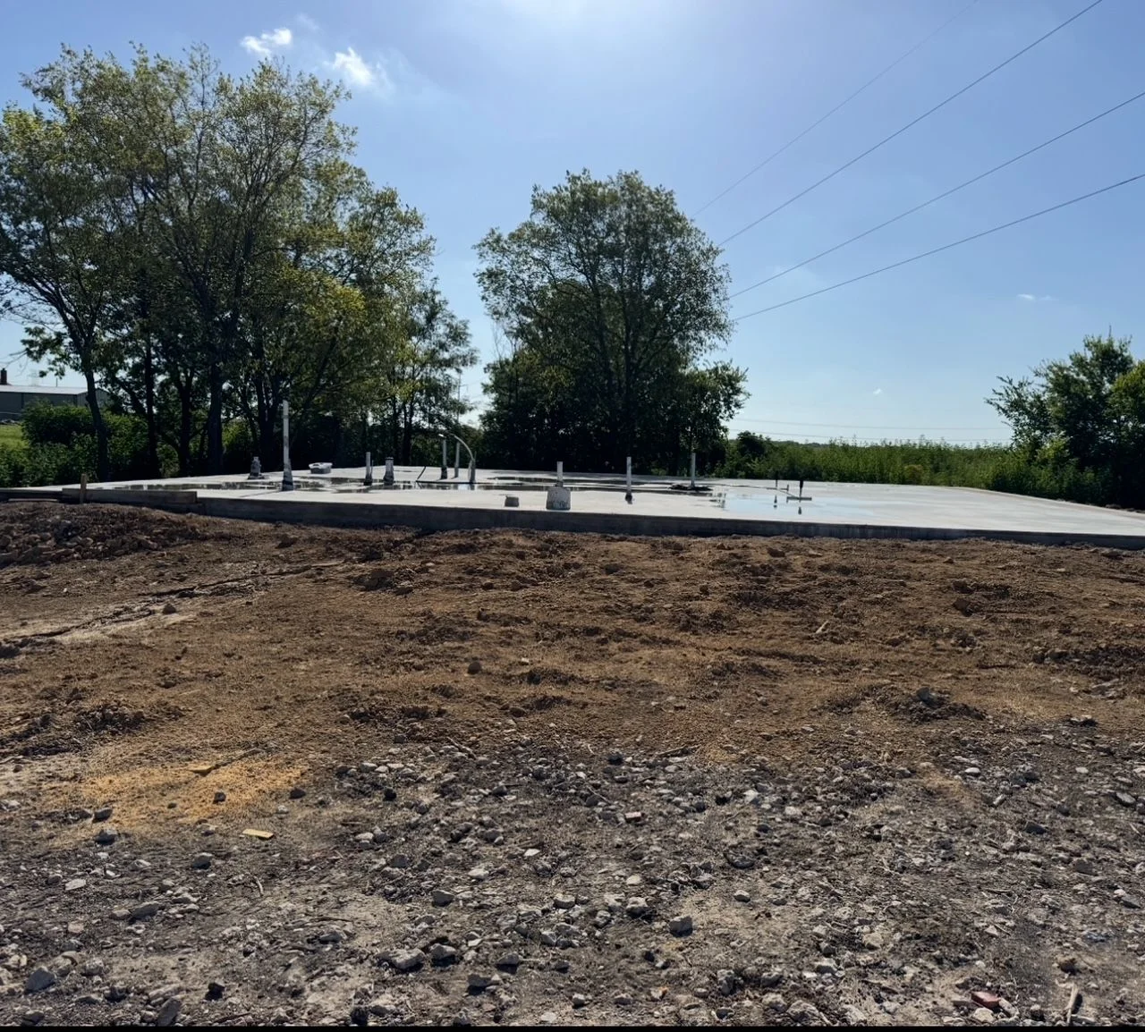 A tracked excavator digging a deep foundation trench in dark expansive clay soil at a residential construction site in Dallas, Texas. The excavated pit shows clean, level walls prepared for a slab foundation pour.