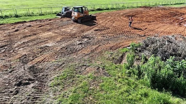 A graded residential lot in Dallas, Texas showing freshly shaped soil sloping away from a concrete foundation with no standing water present. Heavy equipment tracks are visible across the compacted clay surface following rough grading work.