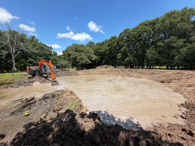 A freshly excavated residential pond on a Dallas property showing shaped basin walls, graded slopes, and compacted clay embankment. An excavator sits at the edge of the site with cleared North Texas terrain in the background.