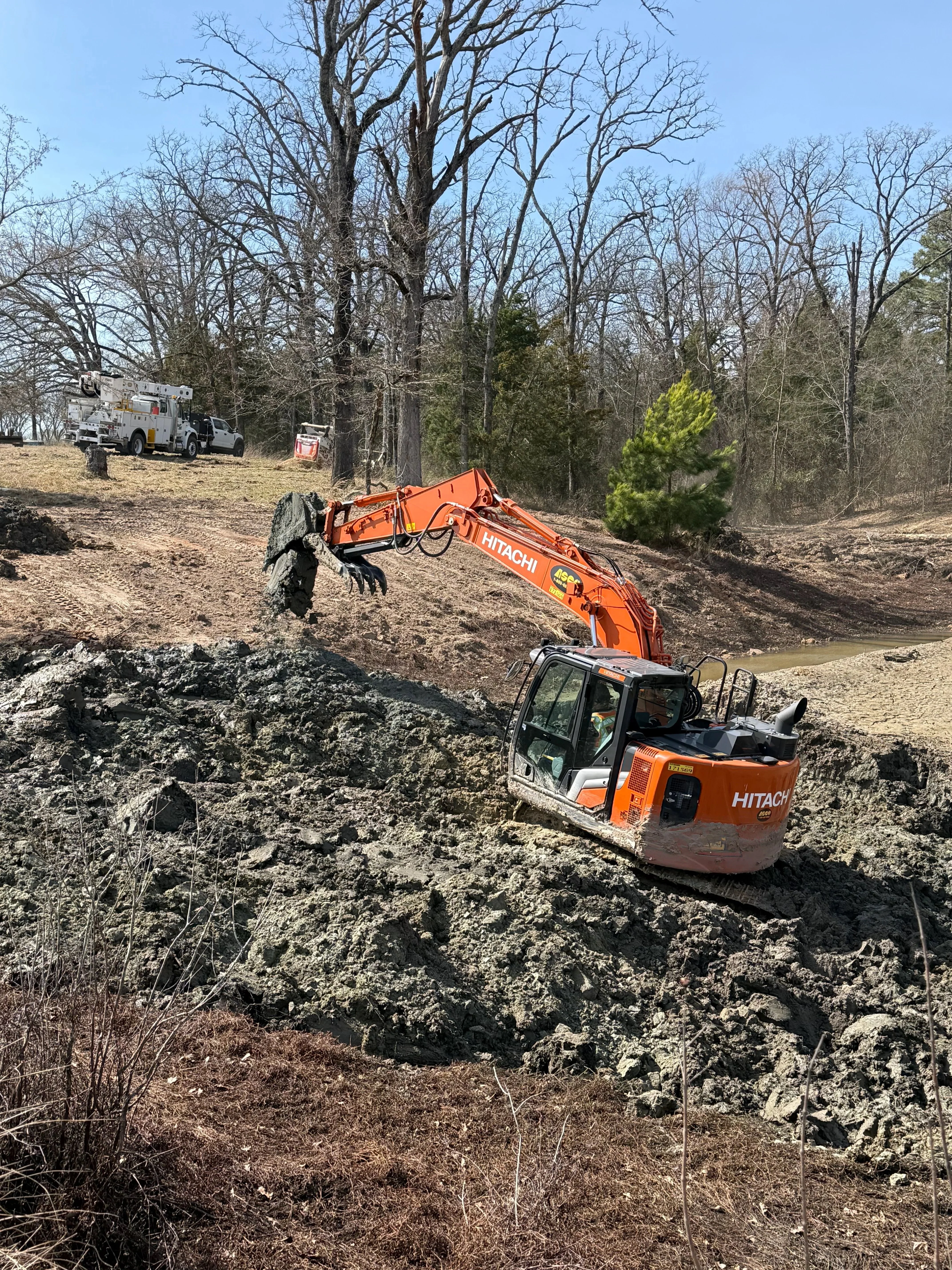 A Dallas construction site drainage channel after sediment removal, showing cleared soil and restored water flow along a graded earthen bank. A worker in safety gear inspects the stabilized channel edge on a North Texas job site.