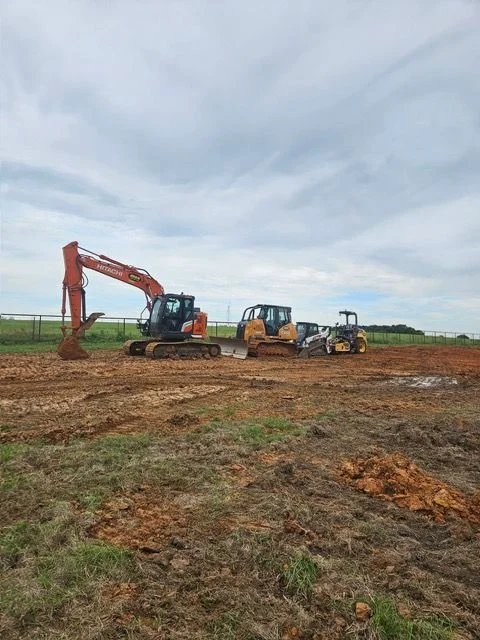 A Dallas residential lot mid-site preparation, showing graded and compacted soil cleared of vegetation and debris. A tracked excavator sits on the leveled ground with a new construction build pad taking shape in the foreground.
